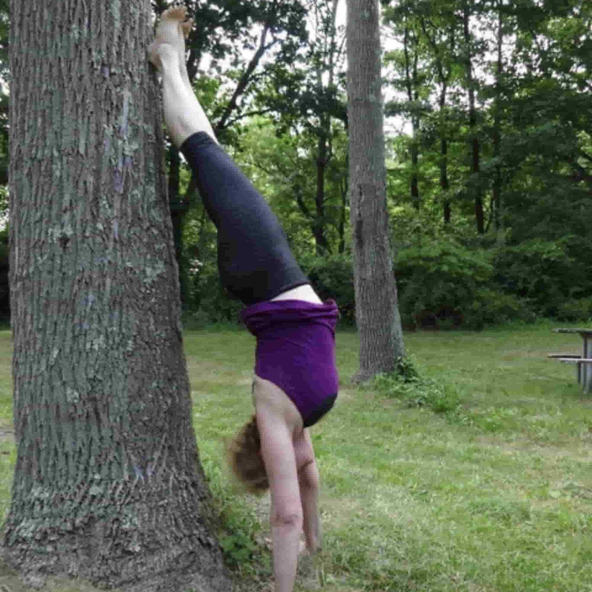 Woman in a purple yoga top and black yoga pants does a handstand against a tree in a green lawn