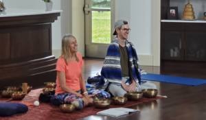 woman in tangerine shirt and man with a blanet sitting with Tibetan singing bowls